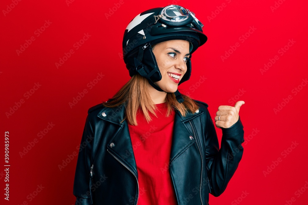 Hispanic young woman wearing motorcycle helmet smiling with happy face looking and pointing to the side with thumb up. Hispanic young woman wearing motorcycle helmet smiling with happy face looking and pointing to the side with thumb up.