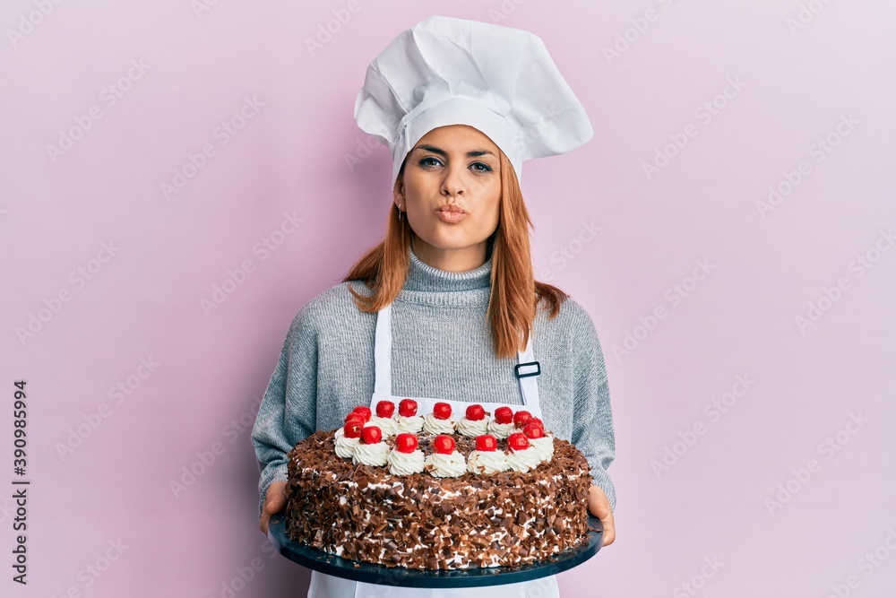 Hispanic young chef woman holding chocolate cake looking at the camera blowing a kiss being lovely and sexy. love expression. Hispanic young chef woman holding chocolate cake looking at the camera blowing a kiss being lovely and sexy. love expression.