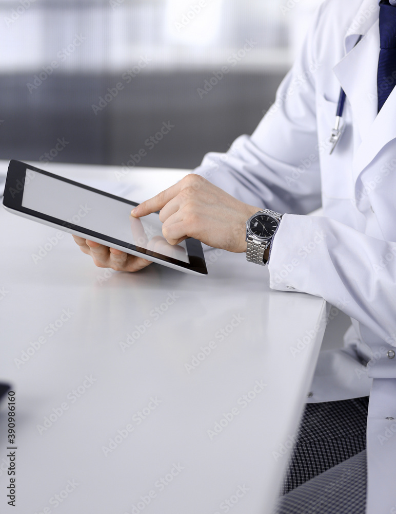 Unknown male doctor sitting and working with tablet computer in clinic at his working place, close-up. Young physician at work. Perfect medical service, medicine concept Unknown male doctor sitting and working with tablet computer in clinic at his working place, close-up. Young physician at work. Perfect medical service, medicine concept
