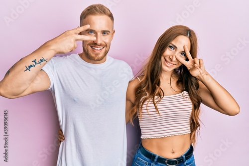 Young couple of girlfriend and boyfriend hugging and standing together doing peace symbol with fingers over face, smiling cheerful showing victory