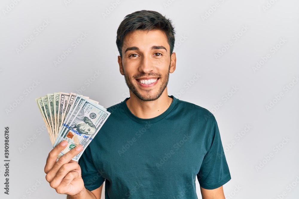 Young handsome man holding dollars looking positive and happy standing and smiling with a confident smile showing teeth Young handsome man holding dollars looking positive and happy standing and smiling with a confident smile showing teeth