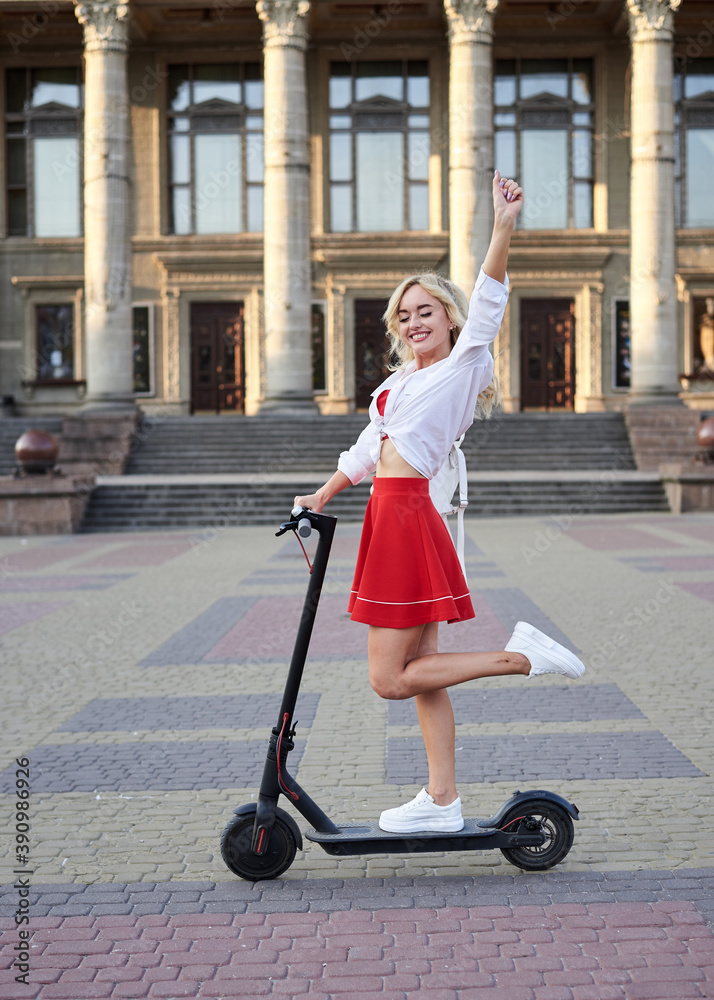 Young blond woman, riding on black electric scooter in city center in front of old historical building with pillars. Summer leisure activity. Spending free time outdoors when weather is warm. Young blond woman, riding on black electric scooter in city center in front of old historical building with pillars. Summer leisure activity. Spending free time outdoors when weather is warm.