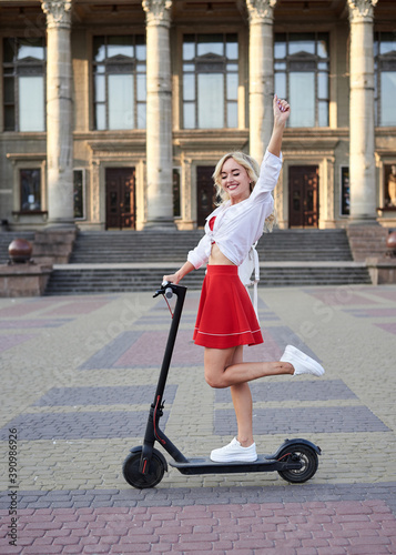 Young blond woman, riding on black electric scooter in city center in front of old historical building with pillars. Summer leisure activity. Spending free time outdoors when weather is warm.