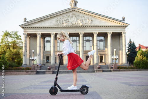 Young blond woman, riding on black electric scooter in city center in front of old historical building with pillars. Summer leisure activity. Spending free time outdoors when weather is warm.