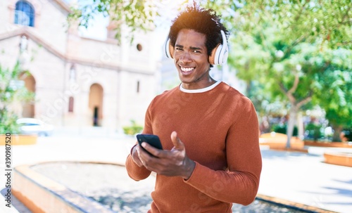 Young handsome african american man wearing headphones. Standing with smile on face using smartphone walking at town street.