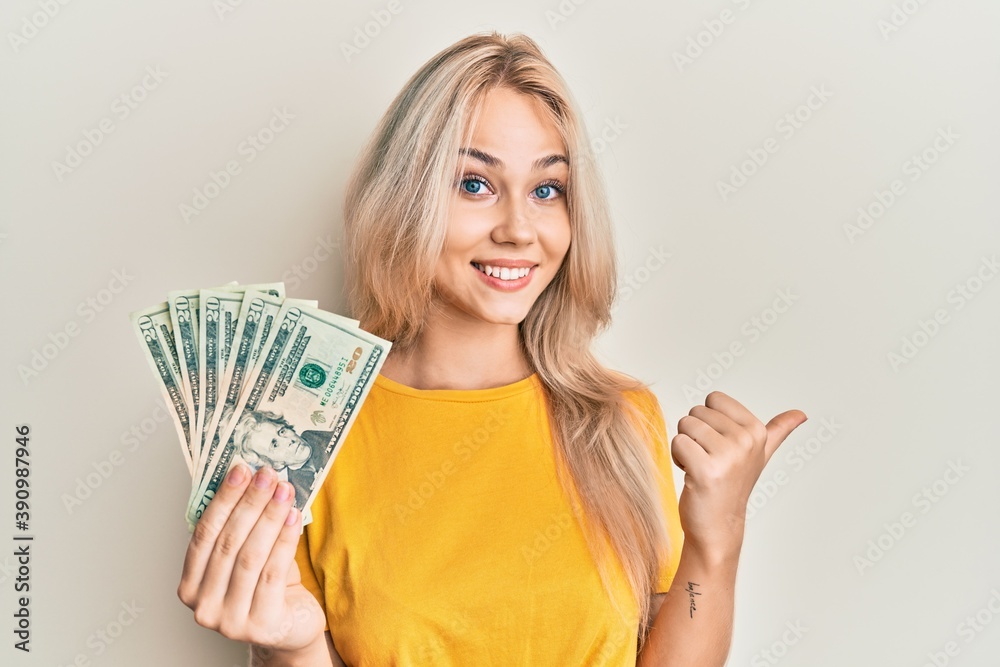 Beautiful caucasian blonde girl holding 20 dollars banknotes pointing thumb up to the side smiling happy with open mouth Beautiful caucasian blonde girl holding 20 dollars banknotes pointing thumb up to the side smiling happy with open mouth