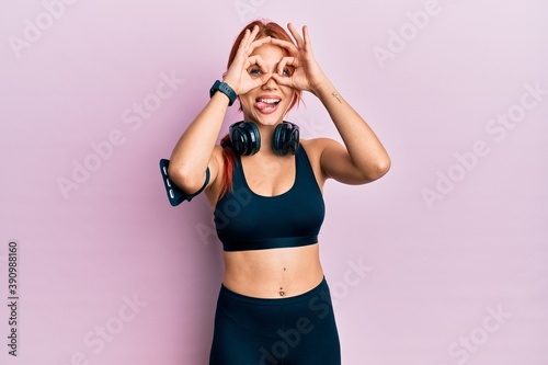 Young beautiful redhead woman wearing gym clothes and using headphones doing ok gesture like binoculars sticking tongue out, eyes looking through fingers. crazy expression.