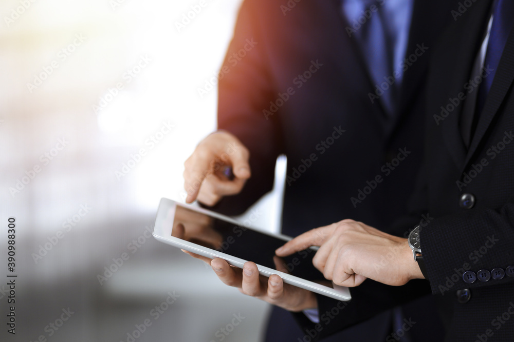 Business people use a tablet computer for discussion of their new project, standing in sunny modern office. Unknown businessman or male entrepreneur with a colleague at workplace