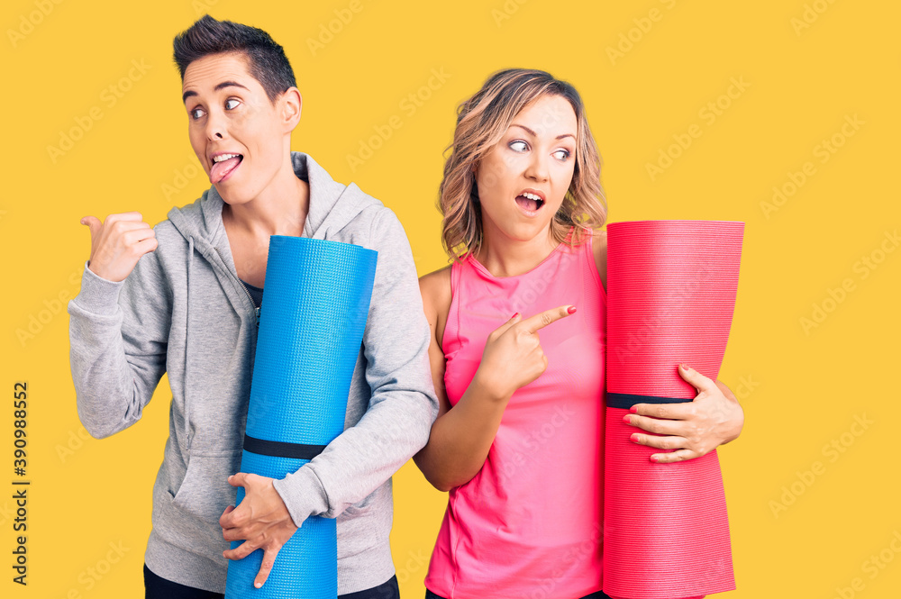 Couple of women holding yoga mat pointing thumb up to the side smiling happy with open mouth Couple of women holding yoga mat pointing thumb up to the side smiling happy with open mouth