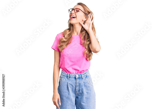 Young beautiful caucasian woman with blond hair wearing casual clothes and glasses smiling with hand over ear listening an hearing to rumor or gossip. deafness concept.