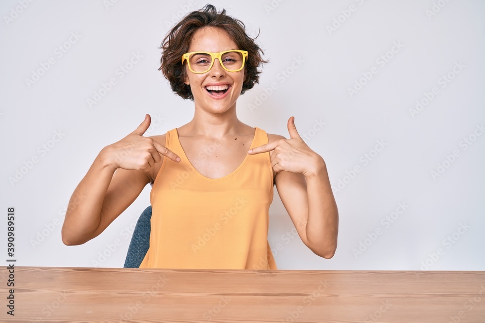 Young hispanic woman wearing casual clothes and glasses sitting on the table looking confident with smile on face, pointing oneself with fingers proud and happy.