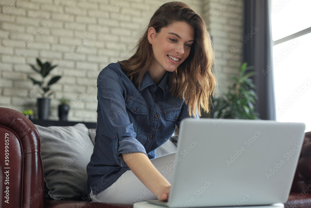 Fototapeta premium Smiling young woman sitting on sofa with laptop computer and chating with friends.