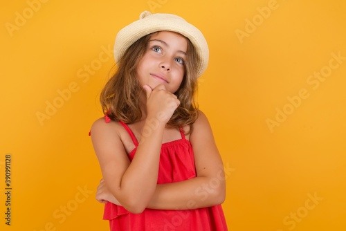 young Caucasian girl wearing red dress and a hat, standing against yellow background smiling friendly holding a hand under chin.