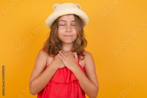 young Caucasian girl standing against yellow backgr closes eyes and keeps hands on chest near heart, expresses sincere emotions, being kind hearted and honest. Body language and real feelings concept.