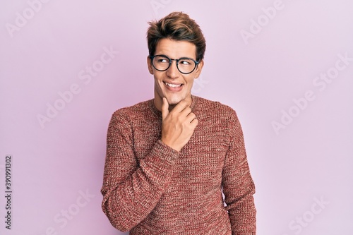 Handsome caucasian man wearing casual sweater and glasses with hand on chin thinking about question, pensive expression. smiling with thoughtful face. doubt concept.