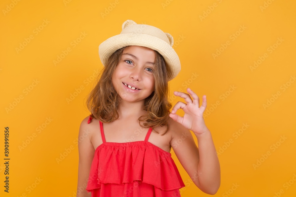 Glad attractive young Caucasian little girl wearing hat and red dress, against orange wall shows ok sign with hand as expresses approval, has cheerful expression, being optimistic. 
