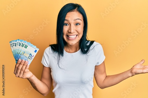 Beautiful hispanic woman holding 20 euros banknotes celebrating achievement with happy smile and winner expression with raised hand