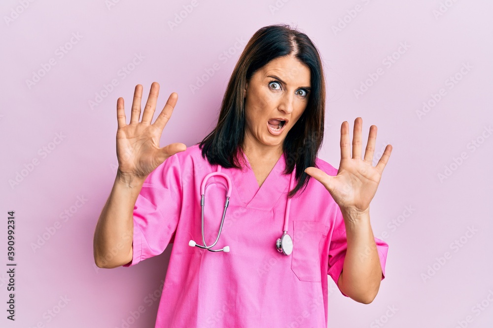 Middle age brunette woman wearing doctor uniform and stethoscope afraid and terrified with fear expression stop gesture with hands, shouting in shock. panic concept. Middle age brunette woman wearing doctor uniform and stethoscope afraid and terrified with fear expression stop gesture with hands, shouting in shock. panic concept.