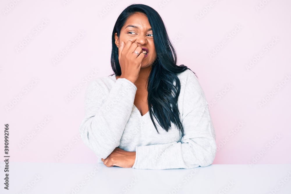 Beautiful latin young woman with long hair wearing casual clothes sitting on the table looking stressed and nervous with hands on mouth biting nails. anxiety problem. Beautiful latin young woman with long hair wearing casual clothes sitting on the table looking stressed and nervous with hands on mouth biting nails. anxiety problem.