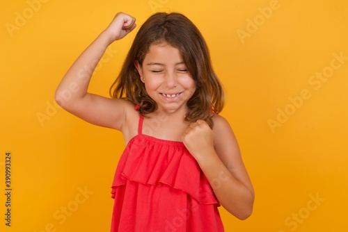 Attractive young Caucasian girl standing against yellow background celebrating a victory punching the air with his fists and a beaming toothy smile.