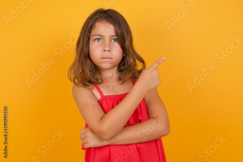 young Caucasian girl standing against yellow background smiling broadly at camera, pointing fingers away, showing something interesting and exciting.