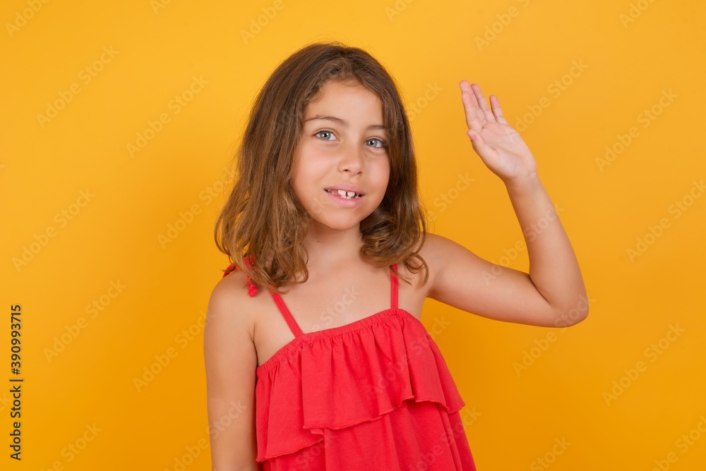 Portrait of cheerful, friendly young Caucasian girl standing against yellow background raise one hand and wave, saying hi or hello as makes goodbye or welcome gesture. Portrait of cheerful, friendly young Caucasian girl standing against yellow background raise one hand and wave, saying hi or hello as makes goodbye or welcome gesture.