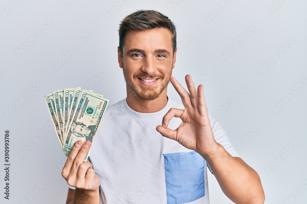 Handsome caucasian man holding 20 dollars banknote doing ok sign with fingers, smiling friendly gesturing excellent symbol Handsome caucasian man holding 20 dollars banknote doing ok sign with fingers, smiling friendly gesturing excellent symbol