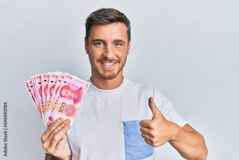 Handsome caucasian man holding 100 yuan chinese banknotes smiling happy and positive, thumb up doing excellent and approval sign Handsome caucasian man holding 100 yuan chinese banknotes smiling happy and positive, thumb up doing excellent and approval sign