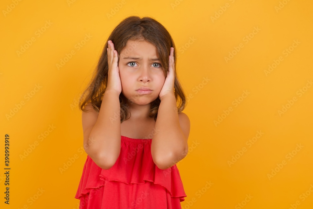young Caucasian girl standing against yellow background holding head in hands with unhappy expression watching sad movie about animals and trying not to cry. young Caucasian girl standing against yellow background holding head in hands with unhappy expression watching sad movie about animals and trying not to cry.