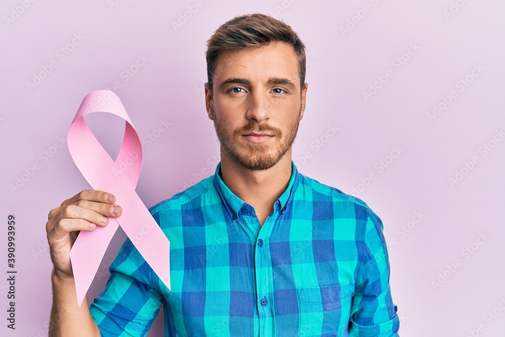 Handsome caucasian man holding pink cancer ribbon thinking attitude and sober expression looking self confident Handsome caucasian man holding pink cancer ribbon thinking attitude and sober expression looking self confident