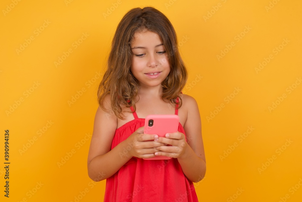 Smiling Caucasian young girl standing against yellow background using cell phone, messaging, being happy to text with friends, looking at smartphone. Modern technologies and communication. Smiling Caucasian young girl standing against yellow background using cell phone, messaging, being happy to text with friends, looking at smartphone. Modern technologies and communication.