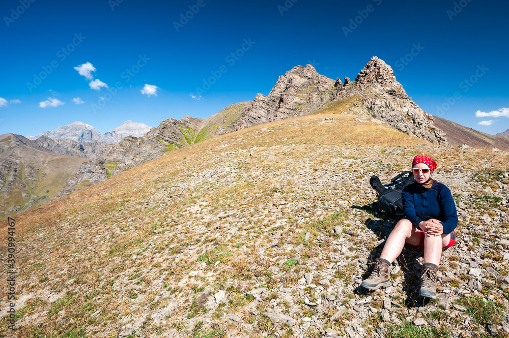 Young trekker woman resting in rocky pass with mountain top in high mountains. Woman with sunglasses sitting next to backpack hiking near Sary Chelek lake Kyrgyzstan, Trekking Central Asia