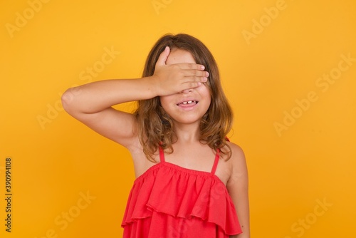 Happy Caucasian young girl standing against yellow background closing eyes with hand going to see surprise prepared by friend standing and smiling in anticipation for something wonderful.