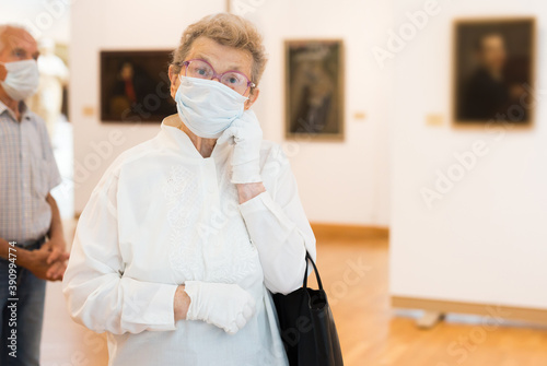 elderly European woman  in mask protecting against covid examines paintings on display in hall of art museum