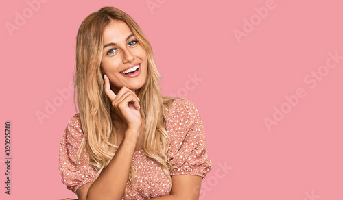 Beautiful blonde young woman wearing summer top looking confident at the camera with smile with crossed arms and hand raised on chin. thinking positive.
