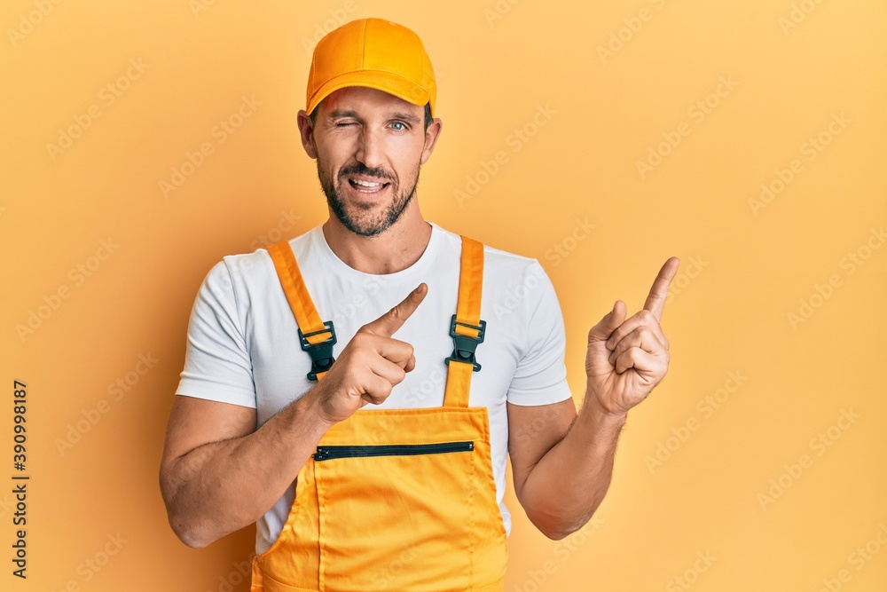 Young handsome man wearing handyman uniform pointing to the side winking looking at the camera with sexy expression, cheerful and happy face. Young handsome man wearing handyman uniform pointing to the side winking looking at the camera with sexy expression, cheerful and happy face.
