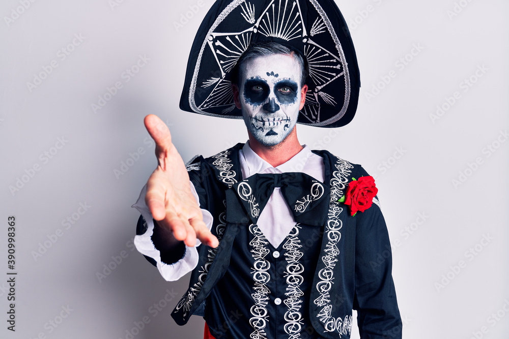 Young man wearing mexican day of the dead costume over white smiling friendly offering handshake as greeting and welcoming. successful business.