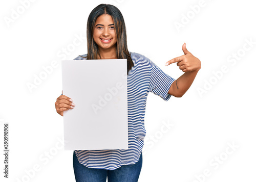 Young latin girl holding blank empty banner smiling happy pointing with hand and finger