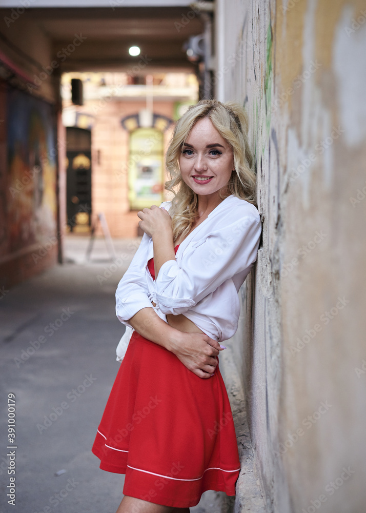 Young blond woman, leaning on old building wall in city center. Three-quarter portrait of female, wearing red and white outfit, posing. Summer leisure activity. Traveler exploring the town. Young blond woman, leaning on old building wall in city center. Three-quarter portrait of female, wearing red and white outfit, posing. Summer leisure activity. Traveler exploring the town.