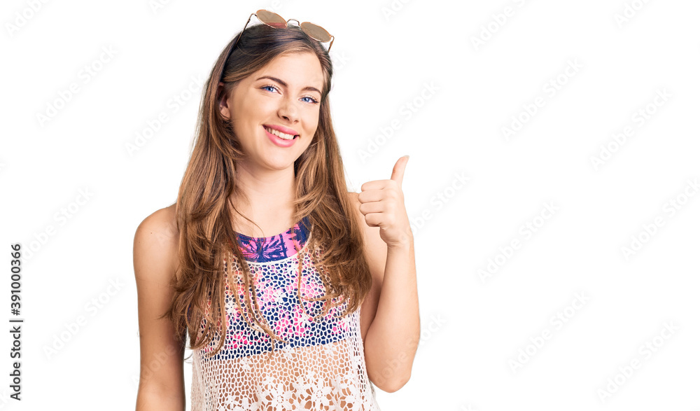 Beautiful caucasian young woman wearing bikini and sunglasses smiling happy and positive, thumb up doing excellent and approval sign Beautiful caucasian young woman wearing bikini and sunglasses smiling happy and positive, thumb up doing excellent and approval sign