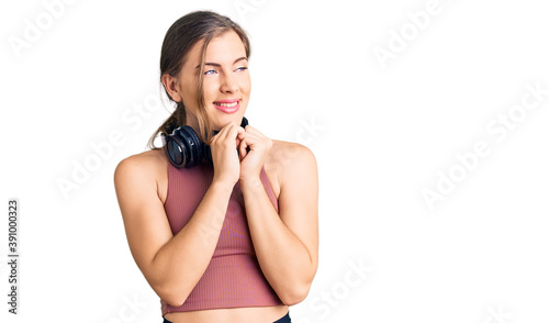 Beautiful caucasian young woman wearing gym clothes and using headphones laughing nervous and excited with hands on chin looking to the side
