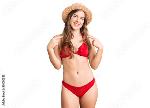 Beautiful caucasian young woman wearing bikini and summer hat looking confident with smile on face, pointing oneself with fingers proud and happy.