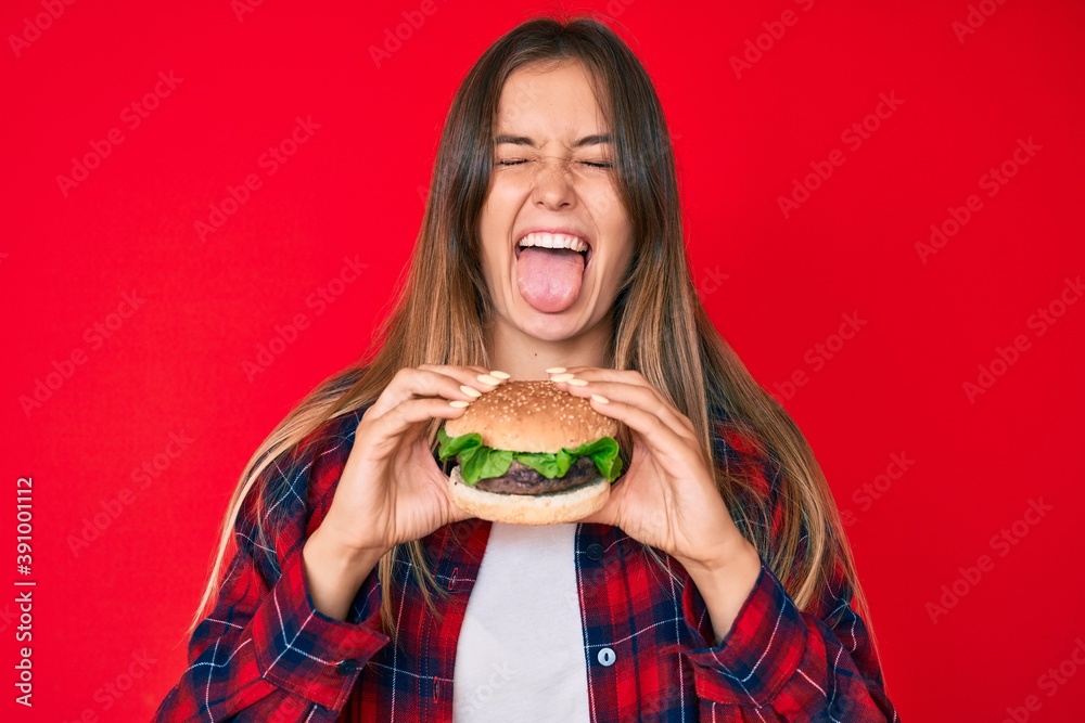 Beautiful caucasian woman eating a tasty classic burger sticking tongue out happy with funny expression. - obrazy, fototapety, plakaty 