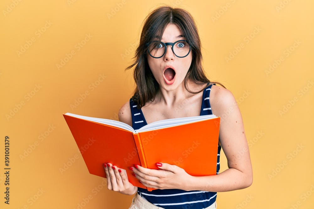 Young beautiful caucasian girl reading a book wearing glasses afraid and shocked with surprise and amazed expression, fear and excited face. Young beautiful caucasian girl reading a book wearing glasses afraid and shocked with surprise and amazed expression, fear and excited face.