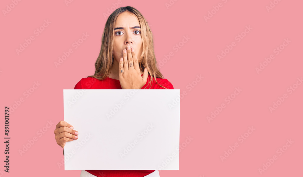 Young beautiful blonde woman holding blank empty banner covering mouth with hand, shocked and afraid for mistake. surprised expression