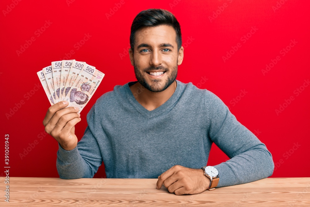Handsome hispanic man holding mexican pesos looking positive and happy standing and smiling with a confident smile showing teeth Handsome hispanic man holding mexican pesos looking positive and happy standing and smiling with a confident smile showing teeth