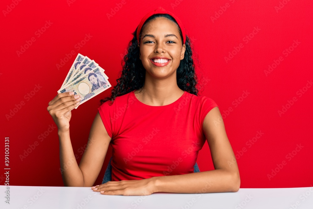 Young african american girl holding japanese yen banknotes sitting on the table looking positive and happy standing and smiling with a confident smile showing teeth Young african american girl holding japanese yen banknotes sitting on the table looking positive and happy standing and smiling with a confident smile showing teeth