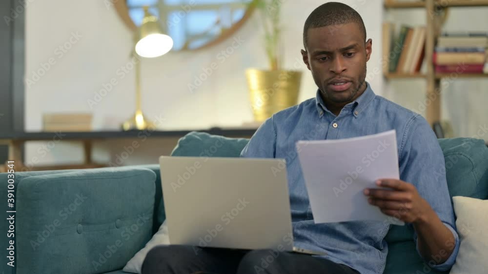 Young African Man with Documents working on Laptop on Sofa Young African Man with Documents working on Laptop on Sofa