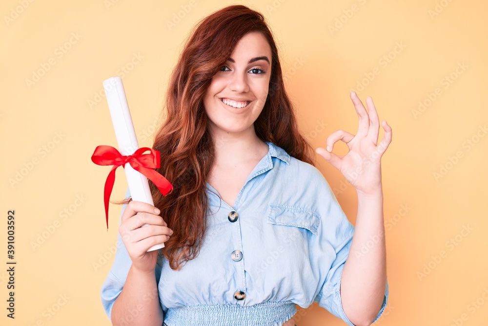 Young beautiful woman holding graduate degree diploma doing ok sign with fingers, smiling friendly gesturing excellent symbol Young beautiful woman holding graduate degree diploma doing ok sign with fingers, smiling friendly gesturing excellent symbol