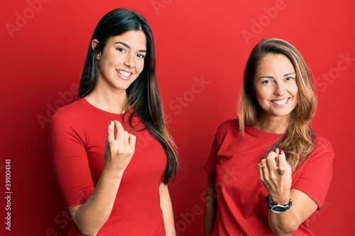 Hispanic family of mother and daughter wearing casual clothes over red background beckoning come here gesture with hand inviting welcoming happy and smiling
